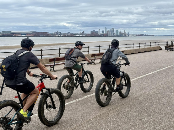 Three cyclists with Liverpool skyline in background across the Mersey