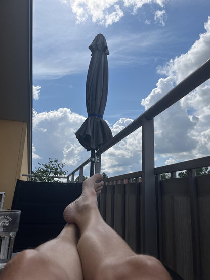 Två bara ben, ur eget perspektiv. Blå himmel kantad av moln. Balkongräcke med ihopfällt parasoll. 