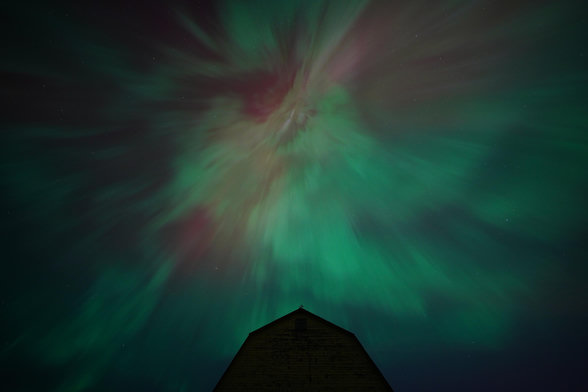 Bright green and red auroras mixed together, but instead of viewing the vertical columns close to the horizon, the vertical columns are directly overhead, so they appear to converge on a point directly above a silhouetted barn. It looks like something out of a horror film.