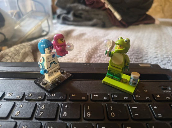 A photo of two minifigs. One is a blue and white android holding up a pink baby in a space suit and the other is a frog suited minifig blowing bubbles. The baby managed to catch a bubble in their hand 🥹