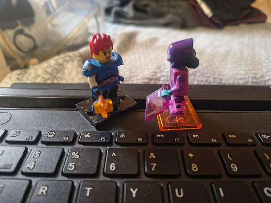 A photo of two minifigs. One is in blue armour with an angry expression and holding a laser chainsaw. The other is a pink cyber spider fig (I think?) playing the role of security officer. They're holding an official looking tablet