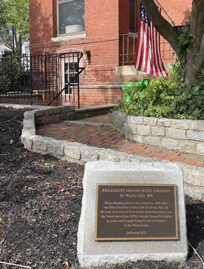 The corner of a brick building with stairs going down to a door set just below griund level. A brick walk going to those stairs curves around a tree at right surrounded by thick, low greenery. A U.S. flag mounted on the building peeks out from behind the tree. Looming large in the foreground, in a dirt flower bed devoid of plantings, is a bronze plaque mounted on a stone slab that reads:
"PRESIDENT OBAMA ROSE GARDEN
At Winter Hill, MA
While attending Harvard Law School (c-1988-1991) our future President lived at 365 Broadway, Apt.1B.
He went on to save us from another great depression, won the Nobel Peace Prize (2009), brought Osama Bin Laden
to justice and brought integrity and intelligence to the White House.
Dedicated 2021"