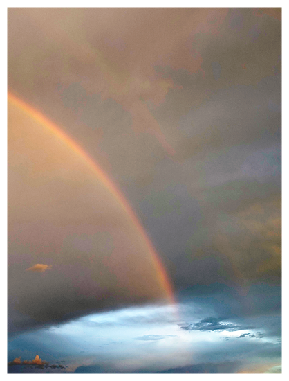 	
A rainbow arches across a dramatic sky, blending with dark clouds above and a bright blue opening below.