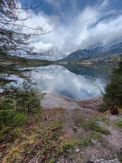 A scene of a blue lake, surrounded by snow topped mountains beneath a cloudy sky. The clouds and sky are reflected in the mirror like surface of the lake