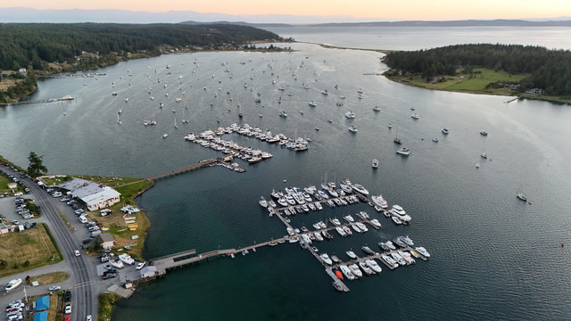 Aerial view looking down into Fisherman Bay from about 400 feet. Two relatively small but completely packed marinas dominate the view. Beyond them are dozens of anchored boats. The bay is small and separated from San Juan Channel by a very narrow strip of land. San Juan Island and the Lompoc Mountains appear in the distance on the horizon where the sky glows with post-sunset peach colored light. 

This photo copyright 2026 by Maria Langer. All rights reserved. Neither this image nor the accompanying alt text may be used to train AI systems.