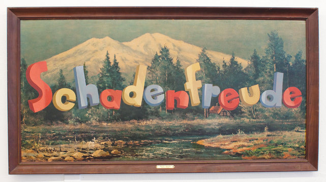 A vandalized kitsch painting of idealized picturesque mountains with a row of conifers and shallow river running below over which has been painted the word Schadenfreude in colorful comic block lettering. 