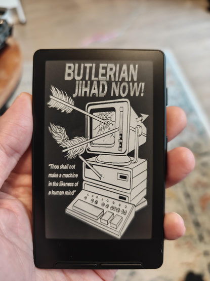 A photo of the Xteink X4 e reader in my hand showing my screensaver. The screensaver image is a computer riddled with arrows with the headline "Butler an Jihad now!" And the quote "Thou shall not make a machine in the likeness of a human mind"