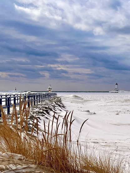 Taken last winter. A view from the shore over a frozen bay on Lake Michigan. On the left a wharf heads out into the bay. In the distance there is a white lighthouse. If the foreground is yellow dune grass blowing in the wind. Roiling clouds of blue and white are overhead. It is very cold and windy.