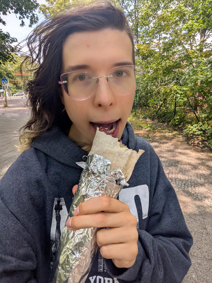 Me in a stolen hoodie making a goofy face eating a Dürum outside :3