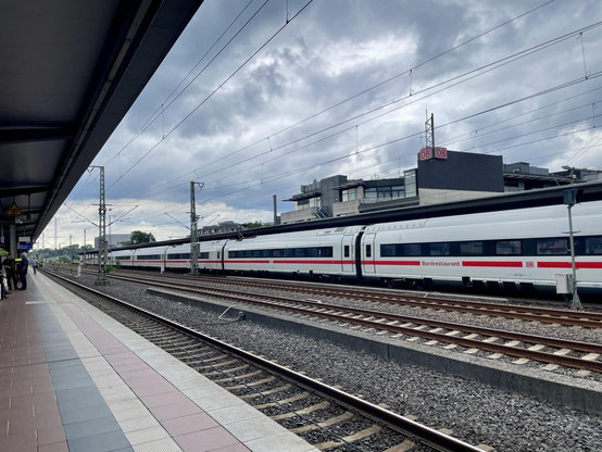 Bahnsteig, Gleise, ein ICE, das Bahnhofsgebäude mit DB-Zeichen unter bewölktem Himmel
