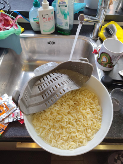 A top-down view shows a large white bowl filled with cooked wavy noodles positioned under a running kitchen faucet. A grey plastic colander with slots sits atop the noodles, through which water from the silver tap flows. flows. In the background, several cleaning bottles sit near the sink. A mug decorated with a colorful cartoon character is visible to the right of the bowl. Small, colorful seasoning packets lie on the edge of the stainless steel sink to the left.