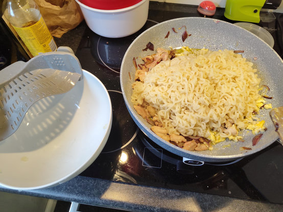 An overhead shot shows various cooking items on a black glass cooktop. In the center, a grey frying pan is filled with wavy noodles, browned pieces of food, and small red flakes. To the left, a white bowl contains a silver metal colander next to a clear plastic bottle filled with yellow liquid. The background includes a white pot with a red rim, a brown paper bag, and various kitchen utensils. The stovetop features circular markings for the heating elements.