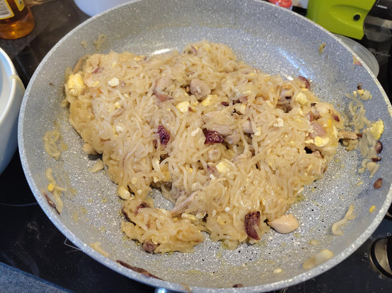 An overhead view shows a grey, speckled frying pan filled with cooked noodles and various ingredients. The translucent, thin noodles are tangled together with small yellow bits, dark reddish-brown pieces, and pale chunks of food. Some residue from the mixture is scattered along the inner sides of the pan. The pan sits on a black stovetop surface. In the blurred background, portions of a white bowl and an amber-colored bottle are visible.