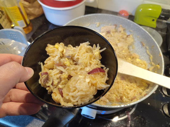 A hand holds a black bowl filled with cooked, pale-yellow noodles mixed with pieces of yellow egg and dark red strips. Beside the bowl sits a large frying pan on an electric stovetop containing more of the same noodle mixture. A white spatula rests in the pan amongst the food. Other kitchen items, including a bottle and a lime green object, are visible in the background. The image is a close-up shot focused on the contents of the bowl and the pan.