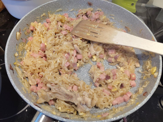 An overhead view of a grey speckled frying pan filled with cooked food. The pan contains small, diced pink pieces, translucent light-colored strands, and small yellow fragments. A wooden spatula rests across the middle of the ingredients. The pan sits on a dark stovetop, with parts of white and green cookware visible in the background.