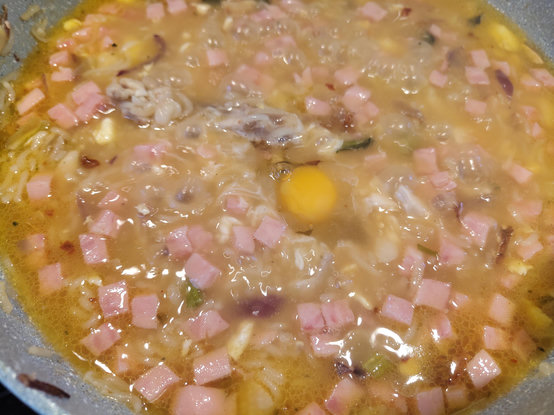 A close-up view shows a pan filled with various ingredients in a translucent, yellowish sauce. Numerous small, pink cubes are scattered throughout the dish. A bright yellow yolk sits near the center, partially surrounded by clear, cooked egg white. Small pieces of darker and lighter-c lighter-colored solids are also visible within the mixture. The liquid appears oily around the edges of the pan.