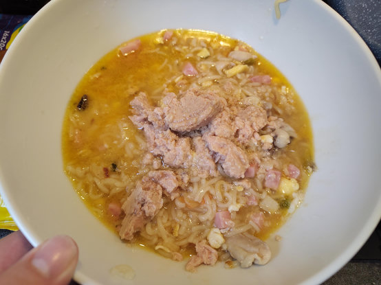 A top-down view shows a white bowl filled with wavy noodles in a yellowish liquid. Large, irregular chunks of light pinkish-tan material are piled in the center on top of the noodles. Small, square pink pieces and tiny white bits are scattered throughout the dish. A portion of a person's thumb is visible at the bottom left corner.