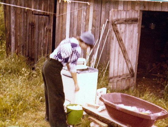 Nuori hoikka nainen, minä parikymppisenä ulkona pesemässä pyykkiä. Vaatetus, tummanvihreät vakosamettihousut, sinivalko-pallokuosinen t-paita ja päässä lierillinen farkkuhattu. 

Vajan rotiskon edustalla penkki nurmikolla. Penkillä pulveripaketti ja pesuharja, sekä punainen, muovinen vauvan amme, jossa pyykkiä huuhtoutumassa. Maassa pulsaattoripesukone ja vihreä ämpäri, jossa siinäkin pyykkiä. 