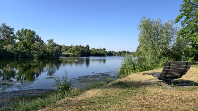 Eine hölzerne, sehr bequeme Bank am ruhigen Ufer eines kleinen Sees mit vielen Bäumen und Sträuchern an den Ufern. Der Himmel ist wolkenlos. Die ruhige Wasseroberfläche spiegelt das Blau des Himmels und die Bäume des gegenüberliegenden Ufers.
A wooden, extra comfortable bench on the shore of a small lake, surrounded by lots of trees and bushes. The sky is clear. The calm water surface is reflecting the blue sky and the trees of the opposite shore.