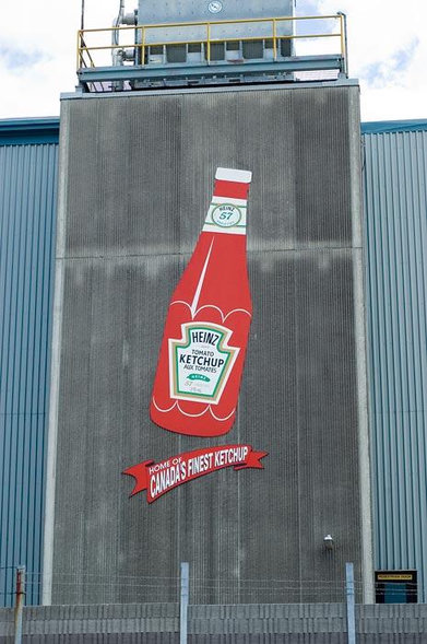 A vertical image of an industrial building. Mounted on a grey wall is an image of a huge, red Heinz ketchup bottle, under which says "Home of Canada's Finest Ketchup."