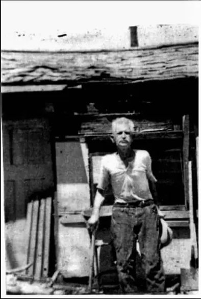 Un retrato fotográfico antiguo en blanco y negro que muestra a William Henry "Burro" Schmidt de pie frente a una estructura rústica.
Persona: Un hombre de edad avanzada con cabello blanco y bigote, vestido con una camisa clara de manga corta y pantalones oscuros desgastados. Sostiene lo que parece ser una herramienta o bastón con su mano derecha.
Entorno: Se encuentra de pie en el exterior, justo delante de una edificación de madera vieja, precaria o de aspecto minero. En el suelo y a su alrededor se aprecian maderas, herramientas y escombros propios de una zona de trabajo rústica.
Estilo: La imagen tiene una calidad granulada, de alto contraste y envejecida, característica de los registros fotográficos de mediados del siglo XX.