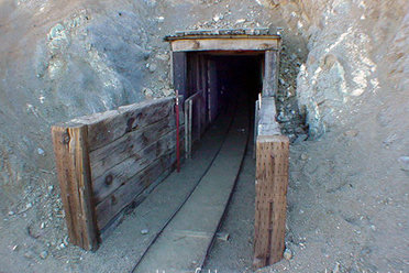 Una fotografía a color de la entrada al túnel de William Henry "Burro" Schmidt, ubicado en una ladera árida y desértica.
La entrada: La boca del túnel está excavada directamente en la roca clara y tierra de la colina. Cuenta con una estructura de soporte rústica de madera vieja y metal corrugado que enmarca la entrada, abriéndose hacia un interior completamente oscuro.
El entorno: El túnel está situado en una pendiente empinada de terreno arenoso y rocoso de color beige o gris claro, salpicada por arbustos secos y vegetación baja desértica. El cielo arriba es de un azul despejado.