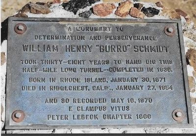 Una fotografía de una placa conmemorativa de metal rectangular, sujeta con grandes tornillos sobre una base de piedra o cemento.
Texto de la placa: Contiene una inscripción grabada en inglés con letras mayúsculas que rinde homenaje a William Henry "Burro" Schmidt. El texto destaca su determinación y perseverancia al cavar a mano un túnel de media milla de largo durante 38 años, completado en 1938. También incluye sus fechas de nacimiento (30 de enero de 1871 en Rhode Island) y fallecimiento (27 de enero de 1954 en Ridgecrest, California), así como la fecha de registro del monumento (16 de mayo de 1970) por la organización E Clampus Vitus, capítulo Peter Lebeck 1866.
Apariencia: La placa de bronce o hierro tiene una pátina envejecida con tonos grisáceos y azulados debido a la exposición a la intemperie.