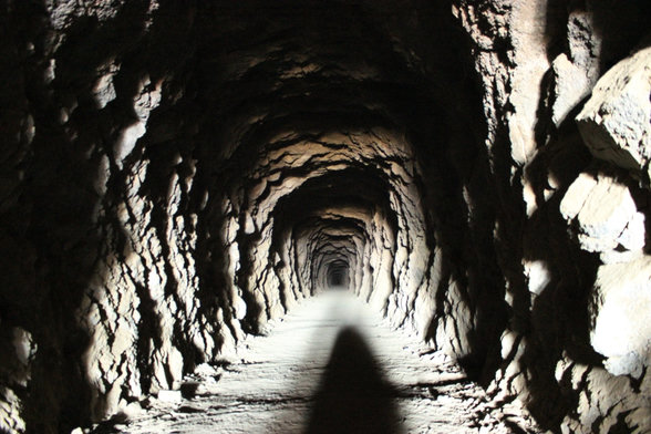 Una fotografía a color tomada desde el interior del túnel de William Henry "Burro" Schmidt, mirando hacia la profundidad del pasaje.
Estructura del túnel: Las paredes y el techo muestran una superficie rugosa e irregular de roca sólida tallada a mano, revelando las marcas del trabajo de excavación. El túnel es estrecho, de forma arqueada y se extiende en línea recta hacia el fondo.
Iluminación: La escena está iluminada artificialmente, posiblemente por la linterna o el flash del fotógrafo, lo que genera fuertes contrastes entre las zonas iluminadas del suelo y los relieves rocosos, y las sombras oscuras en los recovecos de las paredes.
Suelo y perspectiva: El suelo es de tierra o roca aplanada y polvorienta, mostrando en primer plano la sombra alargada de quien toma la fotografía. La perspectiva crea un efecto de profundidad que dirige la mirada hacia el punto más lejano y oscuro del túnel.