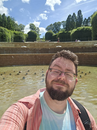 Bearded man taking a selfie (me) in front of a large water feature with very tall hedges in the background. He almost has his eyes closed as it's very bright in the sun