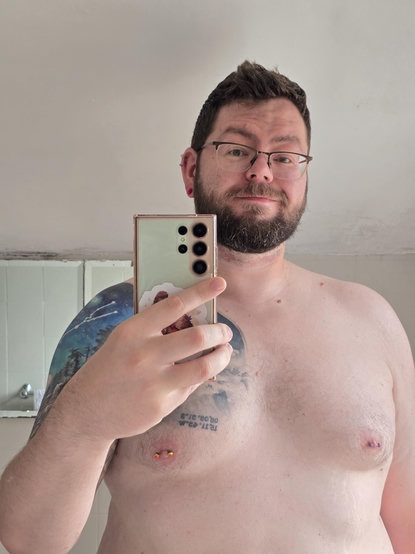Bearded man smiling and taking a selfie in the bathroom mirror (it's me again). He's shirtless with pierced nipples and a tattoo on his chest that continues onto his left shoulder