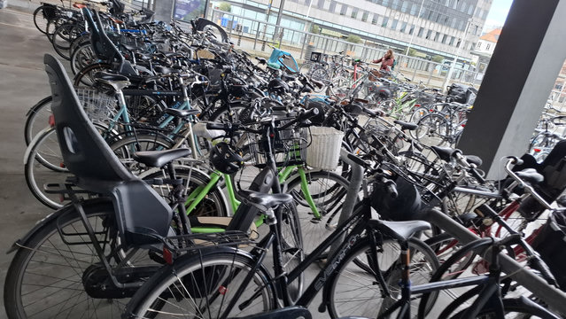 Between the library entrance and the tram stop, a bike park. Rows of bike racks you can place the front wheel in, but the wheel slots are too close together for lots of modern bikes now used for functional everyday transport. We see a tangle of bikes, many with big wide baskets, child seats, pannier bags, wide handlebars, covered trailers, cargo carriers.