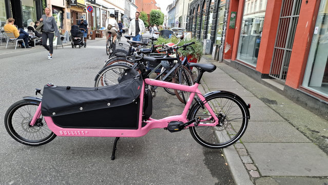 An Aarhus mid-city street, shops and cafes on either side. Pedestrians and cyclists have priority ahead of cars and there’s a 15-kilometre speed limit. We see café tables on the narrow pavement and cyclists, pedestrians and people pushing prams on the road. In front of us, by the kerb on the road, a row of parked bikes. The nearest one is a fabulous, bright pink Bullitt cargo bike with a long front carrier. It’s about two and a half metres long.