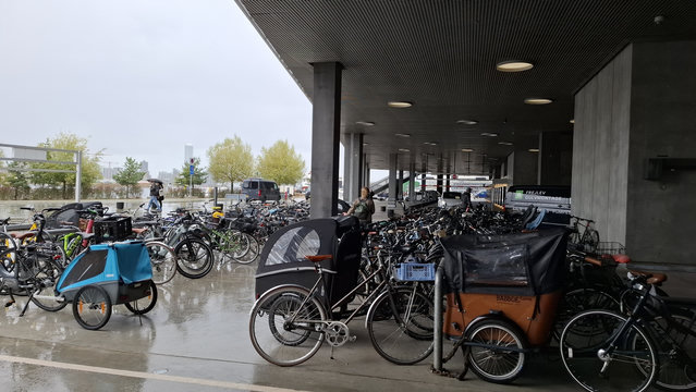 By the harbour, near the library, a bike park in the rain, full of bikes too long and wide for the densely packed bike rack wheel slots. We see bikes with big wide baskets, child seats, pannier bags, wide handlebars, covered front trailers big enough for two adults or four children, cargo carriers.