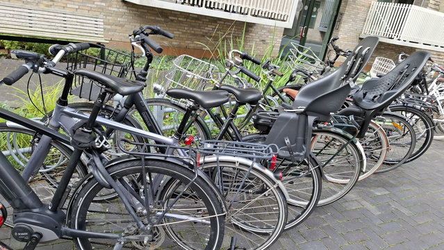 Outside apartments, mid-city Aarhus. Close-up of bike racks. The wheel slots are too close together. They are fine for slim racing bikes. But too narrow for bikes like the ones we see lined up here with wide handlebars, child seats with side foot supports and big wide front baskets. Often, every second wheel slot is left empty and the overflow bikes just get left standing free-style until they blow over in wind.