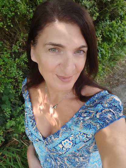 A selfie of an older brunette woman in a gorgeous low cut blue and gold dress in dappled lighting.