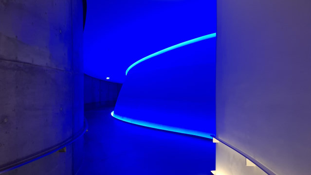 Short version: curving corridor with blue light. Fuller description: underground corridor with concrete floor and roof and concrete walls, curving ahead. The nearest walls are pale grey. A section of wall ahead glows with deep blue light and trimmed in white-green light at the edges. This is the corridor leading to the main chamber for viewing sunrise and sunset through a small portal in the top of the underground Skyspace dome ‘As Seen Below’ designed by James Turrell. Part of ARoS Art Museum. I assume, but do not know, that the curving shapes and light here 16 metres below the earth echo the shapes and curves of Olafur Eliasson's Rainbow Walkway panorama installation just outside, on the top of ARoS museum, but 50 metres up in the air.