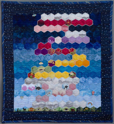 "Morning Mood". Rectangular wall quilt with hexagonal tiles laid out to resemble colourful clouds against a blue sky. Some lapel pins dot the background.