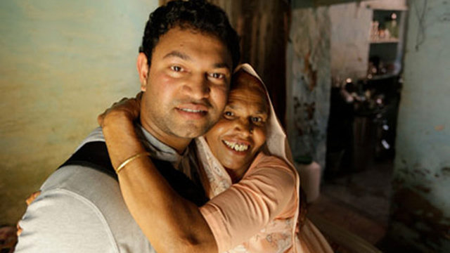 Una fotografía a color de plano medio que muestra a Saroo Brierley (nacido como Sheru Munshi Khan) abrazando afectuosamente a su madre biológica, Kamla Munshi, tras su reencuentro.
Saroo Brierley (Izquierda): Un hombre joven con el cabello corto y oscuro, que viste una camiseta gris claro. Mira directamente a la cámara con una sonrisa leve y serena mientras sostiene a su madre.
Kamla Munshi (Derecha): Una mujer con un velo o bindi tradicional claro sobre la cabeza y vistiendo una prenda de color salmón o rosa pálido. Abraza a su hijo pasando su brazo por el cuello de él y sonríe con gran emoción y alegría hacia la cámara, mostrando los dientes.
Entorno: Se encuentran en el interior de una vivienda humilde con paredes desgastadas de tono azul claro o verdoso. Al fondo, a la derecha, se observa una apertura que da hacia otra habitación más oscura de la casa.