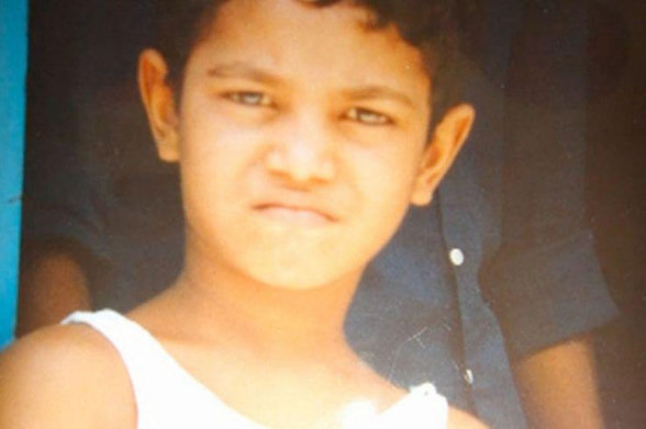 Un primer plano fotográfico a color de un niño pequeño, identificado como Saroo Brierley en su infancia.
Rostro y expresión: El niño mira fijamente hacia la cámara con una expresión seria y el ceño ligeramente fruncido. Tiene el cabello oscuro y corto.
Atuendo: Lleva una camiseta blanca de tirantes.
Fondo: Detrás de él se observa de manera difuminada el torso de otra persona vestida con una camisa oscura, y a la izquierda se aprecia un fragmento de una superficie o pared de color azul brillante.