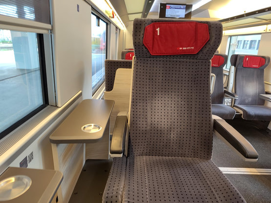 A picture of the interior of a train carriage. The camera is pointed at an opposite seat, with no tray table available. The seat cushion is red, showing the SBB logo and a first class sign. In contrast to Deutsche Bahn first class, the seat is appointed in soft fabric, not leather. 