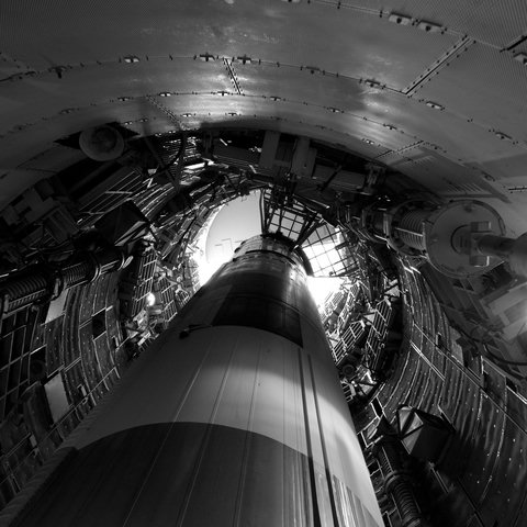 Looking up at a missile pointed upward inside a cramped underground silo, which is lined with retractable platforms and various apparatus.