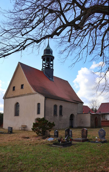 A small village church in Klein Glien with light-coloured rendered walls and a red terracotta tiled gable roof, featuring an integrated bell tower with a pointed top. The church has arched windows, small square windows near the roof, and a main arched entrance. In the foreground is a churchyard cemetery with numerous gravestones and tombstones of varying sizes and designs. A bare deciduous tree with spreading branches frames the top of the scene, while a small conifer and a wooden bench sit near the church. The sky is blue with white clouds, and other red-roofed buildings are visible in the background.