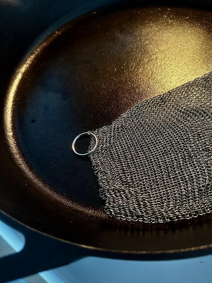 A cast iron skillet is gleaming after cleaning with the metal mesh cloth shown in the pan. It’s early morning so the light is favorable for atmospheric beauty. Image is most of the black pan and handle going to the left bottom corner. Mesh is to the right and center, with a small ring for hanging. Above it to the right is a warm yellow gleam of light reflecting on the surface of a clean plan with oiled surface. 