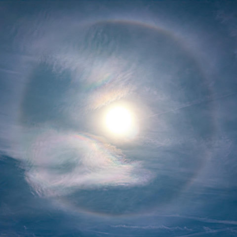 A picture of the Sun high in the sky, with some light cloud. The Sun is ringed by a colourful halo with a diameter of 22º, while there are also colours in the clouds inside the ring thanks to iridescence.