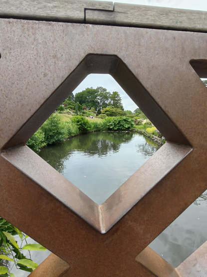Looking through the diamond-shaped space on a metal bridge, beyond is gently rippling water, green bushes, and a hilly rock garden surrounded by tall trees.