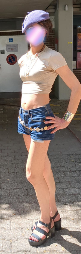 Picture of Nora pausing in the sun in her croptop and minishorts. She has a lilac beret, short hairs, a lot of collars hanging around her neck. The croptop is cream the shape suggesting neat boobies, she wears are minishort in denim, with a shiny loosely tied metal belt, she wear a very reflective tin bracelet, she rests her hand on her short. Her legs are long and bare, supported by plateformer-black sandal clad feet.