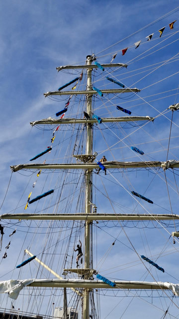 Different ships and angles, same general scene – same alt text as other photos follows; Short version: Sailors on masts. Fuller descriptions: Young sailors dotted around high up on the ropes and riggings and masts of a ship, working on the sails while moored at the Tall Ships Race in Aarhus Denmark (last week). Deep bright blue sky. Coloured bunting on the ships.
