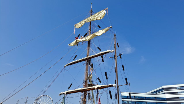 Short version: Sailors on masts. Fuller descriptions: Young sailors dotted around high up on the ropes and cables and riggings and masts of a ship, working on the sails while moored at the Tall Ships Race in Aarhus Denmark (last week). Deep bright blue sky. Coloured bunting on the ships.