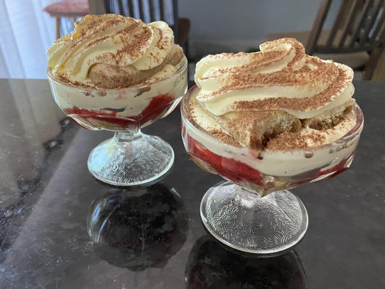 Two stemmed dessert cups with layers of strawberry and cream and sprinkled brown powder on top. 