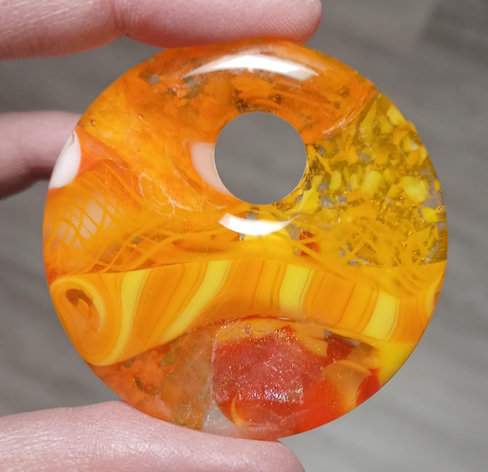 round pendant with hole in yellows, oranges, reds and clear. there's just a bit of dicroic glass along the bottom and there is a part of the design that you can tell is a spiral thing laid flat because you can see the twist where i cut it at the end